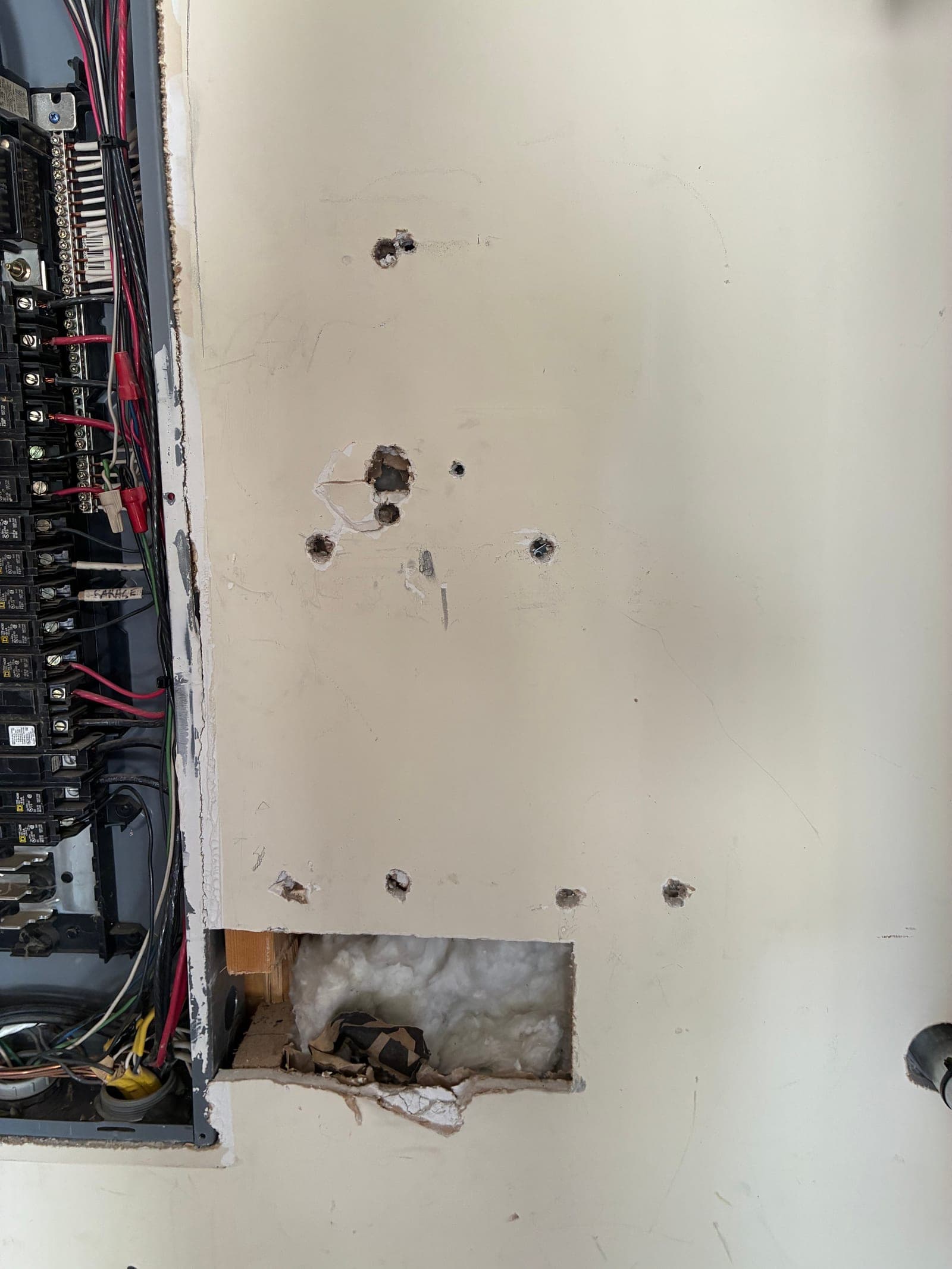 Drywall with several holes before repair, next to an open electrical panel